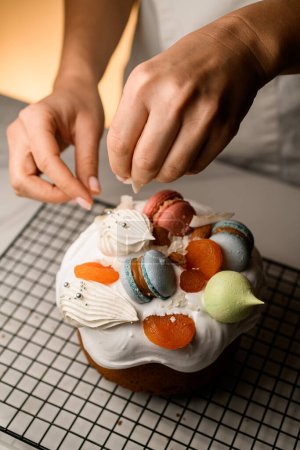 Bakers hands decor beautiful Easter cake with white glaze colorful macaroons, dried apricots and meringue of various shapes