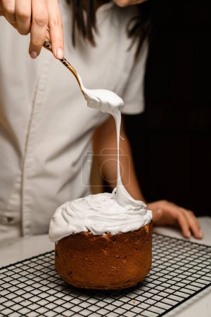 Baker holds a spoon in her hand, from which white cream flows onto the Easter cake standing on a metal grid