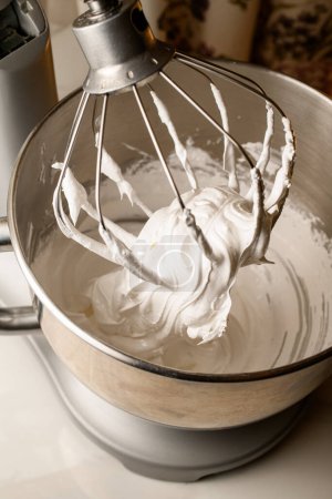 Standmixer mit einem großen Metallbesen schlägt weiße Buttercreme in einer runden tiefen Schüssel auf einem weißen Küchentisch