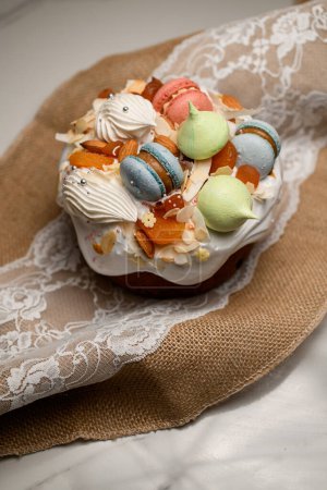 Beautiful and tasty Easter cake decorated with white icing, colorful beret macaroons of different shapes and other confectionery decor stands on a towel with lace