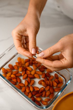 Brown almonds lie in a rectangular glass form on white table, female hands hold peeled almonds