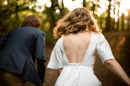 Téléchargez les photos : Homme et sa femme, vêtus d'une robe à dos ouvert, marchent dans la forêt, se tenant la main, s'éloignant de la caméra - en image libre de droit