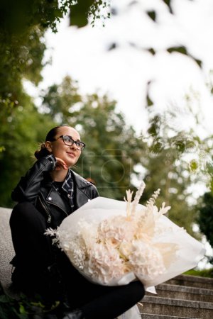 Young pretty girl is holding a huge bouquet of white hydrangeas and white roses on her lap, sitting on the concrete railing of the steps against a blurred background of green trees