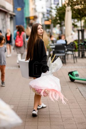 Hübsche junge Frau hält einen riesigen Strauß weißer Hortensien und Rosen in die Höhe, während sie den Bürgersteig hinuntergeht und sich der Kamera entgegendreht.