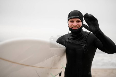 Happy smiling male surfer standing on seashore in wet clothes, water drops on face on blurred seascape background