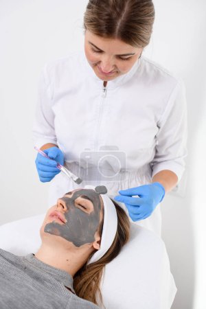 Kosmetikerin Frau in weißen Overalls und blauen Gummihandschuhen fertigt mit Hilfe kosmetischer Werkzeuge eine Salon-Gesichtsmaske für ein Mädchen