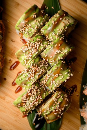 Vue du dessus des sushis enveloppés dans du vert décoré de sésame blanc et d'une épaisse sauce marron clair sur une longue feuille verte sur une planche de bois