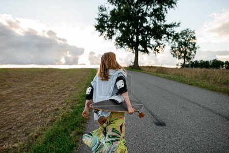 In a dynamic scene, she showcases movement, posing with a longboard on a country road near a tree.