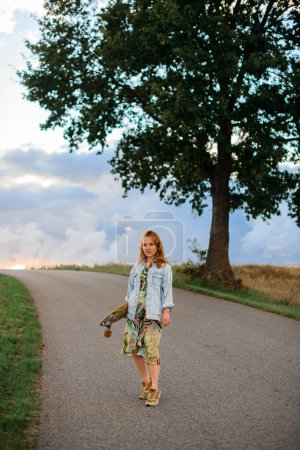 Along a country road, she adds a touch of style, wearing a light blue denim jacket and posing with a longboard near a tree.