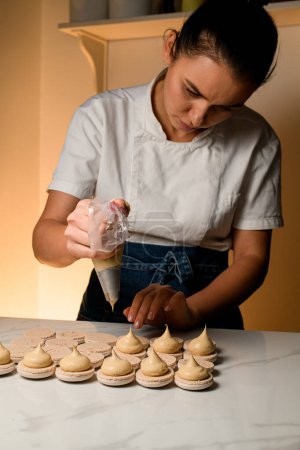 Mains professionnelles d'un spécialiste culinaire fabriquant un délicieux macaron beige clair sur une table en marbre à l'aide d'un sac à pâtisserie