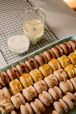 Many macarons in neutral natural color with creamy filling in plastic container and grill grate on a table