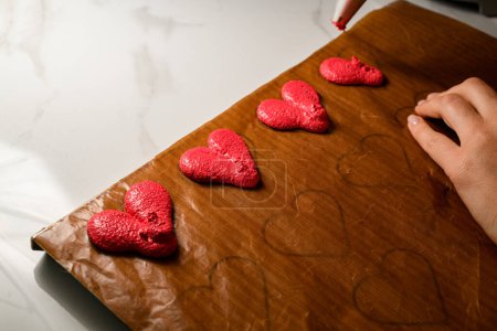 Zubereitungen für Kuchen in Herzform in roter Farbe auf Pergament und Frauenhände mit heller Haut