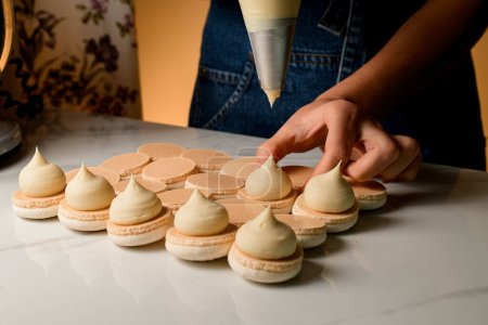 Chef pâtissier professionnel fait le remplissage de macaron avec de la crème marron clair à l'aide d'un sac de pâtisserie brun clair