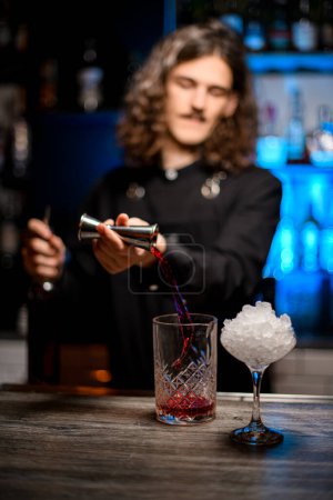 Focus on mixing glass into which bartender pours wine from jigger, tall stemmed glass filled with crushed ice