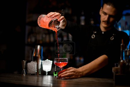 Bartender holds a cocktail glass high and pours the drink into a stemmed glass, straining the liquid through a bar strainer
