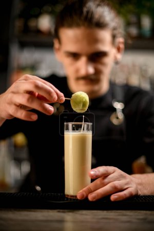 Focus on a tall glass filled with a white cocktail, from which the bartender pulls a soaked corn ball on a skewer, which drips into the glass