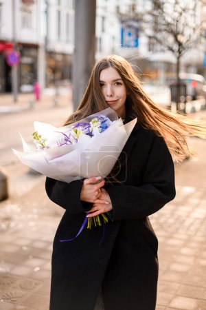 Wonderful young girl with brown long hair in black coat standing on a city street with voluminous bouquet of flowers