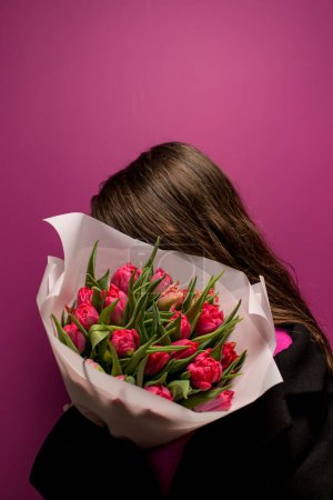 Rostro oculto de niña inocente en un paño negro con un hermoso ramo grande de tulipanes rojos sobre un fondo púrpura
