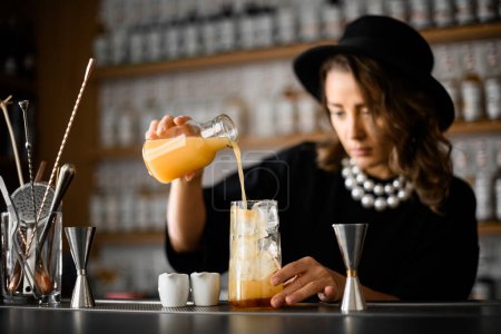 Female bartender pours juice from a jar into a tall glass with ice cubes, focus on the foreground