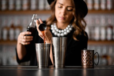 Female bartender pours a brown cocktail drink from a jigger into a shaker standing on a bar counter next to a metal mug with ice, holding a bottle in the other hand