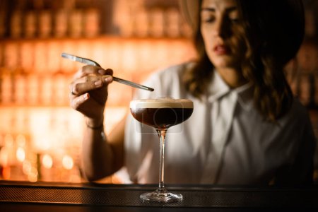 Tall glass with a brown cocktail with foam stands on the bar counter, over which a female bartender holds a pair of tweezers on a blurred background