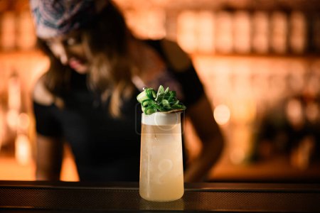 Focus on a tall glass with an iced cocktail, decorated on top with greenery as decorations, on a blurred background where a bartender is standing