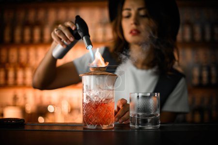 Barkeeperin richtet einen brennenden Gasbrenner auf einen Keramikdeckel mit einer Flamme, die ein Glas bedeckt, um einen Cocktail mit Eis und einem braunen Getränk zu mixen