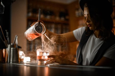 Woman bartender in a white t-shirt and vest pours a cocktail with ice from a mixing glass into another glass, straining the drink through a sieve, smoke spreads near the glasses