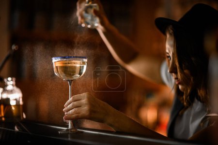 Glass with a tall metal stem and a blue rim sits on the bar, filled with a bubbly champagne-colored cocktail as a female bartender sprays it