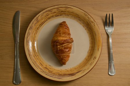 Photo for View from above of an isolated croissant on a plate next to silver cutlery, fork and knife on a plank table - Royalty Free Image