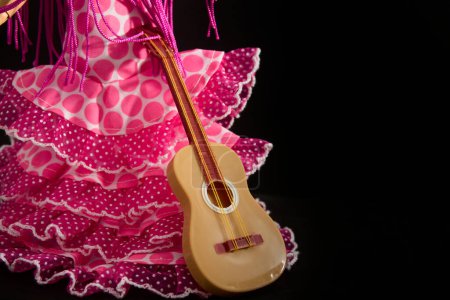 Imagen de la cola de un disfraz de faralaes para bailar flamenco junto a una guitarra todo sobre un fondo negro con espacio para copiar
