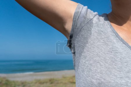Téléchargez les photos : Gros plan d'une femme avec sa main levée dans un T-shirt gris sur lequel la sueur est visible par une journée d'été ensoleillée en mer - en image libre de droit