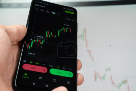 Nahaufnahme der Hand eines Mannes mit einem Smartphone mittels der Analyseanwendung zur Analyse des Finanzmarkts von Kryptowährungen, einer Grafik des Investitionswachstums auf dem Hintergrund eines Monitors