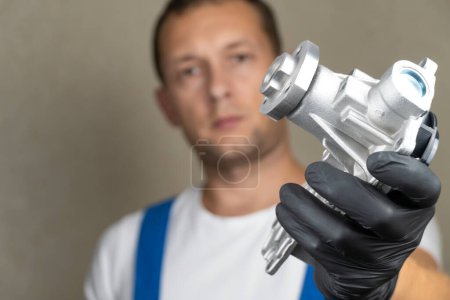 Photo for Car mechanic in blue jumpsuit holds water pump for cooling in his hands and rotates it in black gloves. . Concept of replacement of pump in engine in car, automotive spare parts. - Royalty Free Image