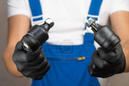 Photo for Auto mechanic in blue jumpsuit holds steering tips or ball bearings of car in his hands and rotates it in black gloves.. Car suspension repair concept, auto parts. - Royalty Free Image