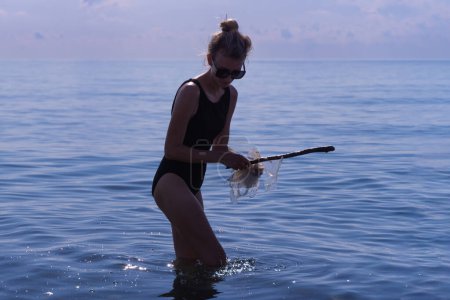 Photo for Young woman in a black one-piece swimsuit with the help of a stick collects floating cellophane bags right into the sea. Ecological catastrophe of the modern world, environmental pollution. - Royalty Free Image