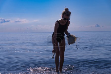 Photo for Young woman in sunglasses, a black one-piece swimsuit with the help of a stick collects floating cellophane bags right into the sea. Environmental pollution, taking care of nature.Ecology of the world - Royalty Free Image