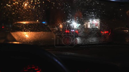 Raindrops on car windshield. Car standing on streets of evening city, blurred background of traffic light and moving cars with street lights.