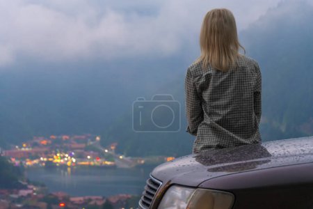 Téléchargez les photos : Jeune femme blonde en chemise à carreaux est assis dans une voiture, admirant la belle vue sur les montagnes et le lac tout en voyageant par une journée nuageuse, l'accent est mis sur la fille. Uzungol, Turquie. - en image libre de droit