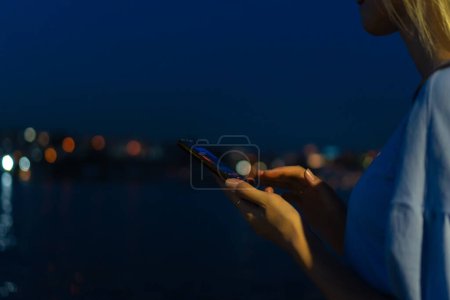 Téléchargez les photos : Gros plan des mains d'une femme avec un téléphone portable sur le fond d'une ville floue avec des lumières, de l'eau, de l'espace de copie. - en image libre de droit