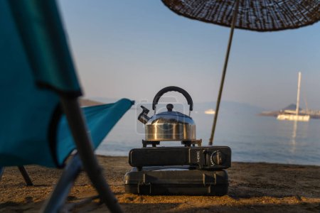 Téléchargez les photos : Bouilloire en métal bouillante sur un poêle à gaz portable sur fond de mer calme, parasol, chaise pliante au lever du soleil. Voyages et loisirs de plein air. - en image libre de droit