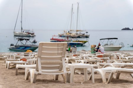 Téléchargez les photos : Gros plan de chaises longues en plastique blanc sur la plage sur fond de mer floue, de montagnes, de yachts et de bateaux. - en image libre de droit
