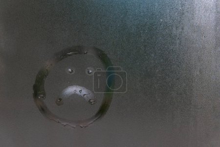 Téléchargez les photos : Triste visage souriant dessiné avec un doigt sur le verre mouillé et brouillard de la pluie. Concept de solitude, tristesse, mauvais temps - en image libre de droit