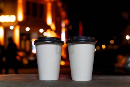 Téléchargez les photos : Gros plan de deux tasses en papier recyclé blanc écologique avec café ou thé debout sur un banc sur le fond d'une ville nocturne floue avec des lumières. Prends le café avec toi.. - en image libre de droit