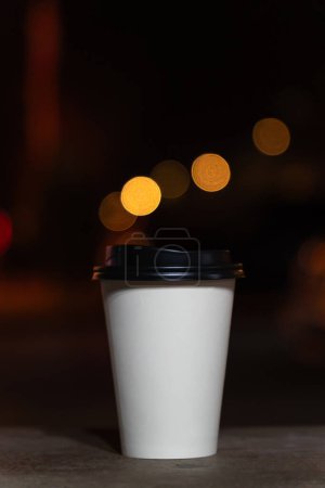 Téléchargez les photos : Gros plan d'une tasse en papier blanc jetable avec café ou thé sur un banc sur le fond d'une ville floue la nuit avec des lumières. Café à emporter. Photo verticale - en image libre de droit