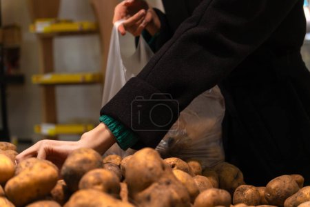Téléchargez les photos : Une femme en manteau noir emballe des pommes de terre dans une épicerie. Acheter des pommes de terre dans le magasin. - en image libre de droit