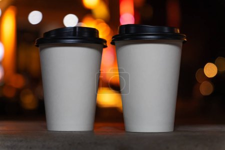 Téléchargez les photos : Gros plan de deux tasses en papier blanc écologique avec café ou thé debout sur un banc sur le fond d'une ville nocturne floue avec des lumières. Prends le café avec toi.. - en image libre de droit