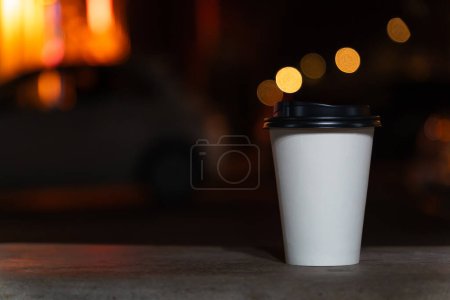 Téléchargez les photos : Gros plan d'une tasse en papier blanc jetable avec café ou thé sur un banc sur le fond d'une ville floue la nuit avec des lumières. Café à emporter - en image libre de droit