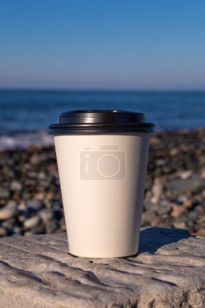 Téléchargez les photos : Gros plan de deux tasses en papier blanc debout sur une pierre au bord de la mer par une journée ensoleillée, copiez l'espace. Café à emporter - en image libre de droit