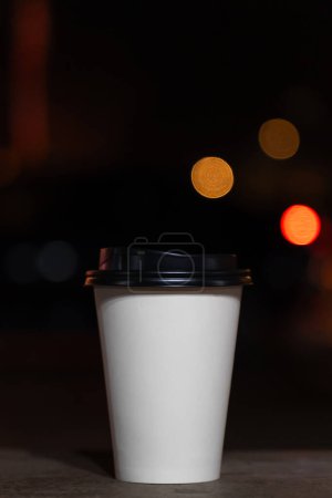Téléchargez les photos : Gros plan d'une tasse en papier blanc jetable avec café ou thé sur un banc sur le fond d'une ville floue la nuit avec des lumières. Café à emporter. Photo verticale - en image libre de droit